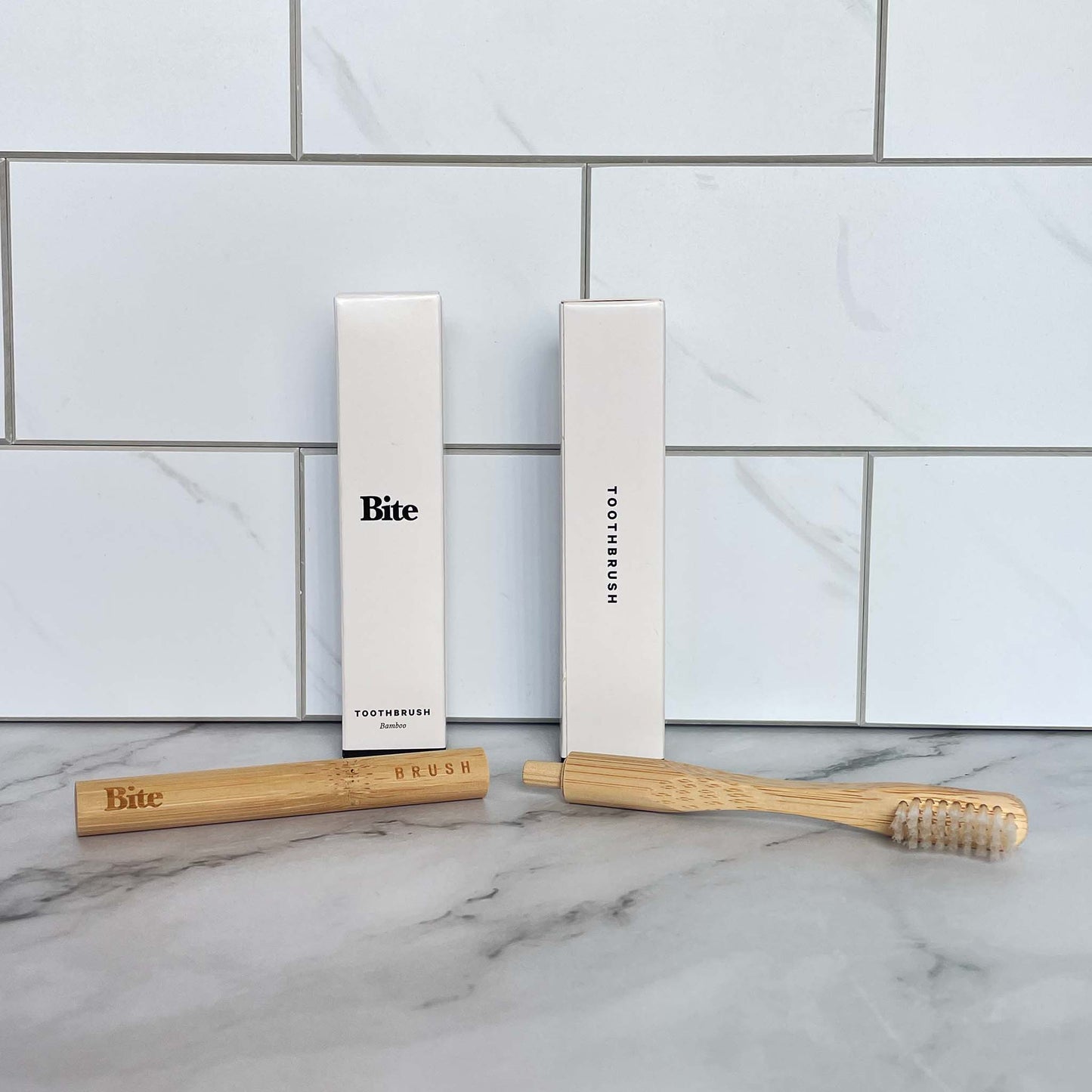 A bamboo toothbrush with packaging (front and side) on a marble surface with tiled wall background