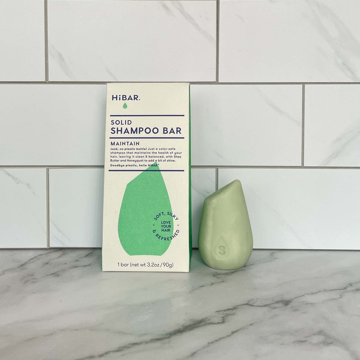 Green solid shampoo bar with HIBAR Maintain Shampoo packaging on a marble surface and tiled wall background