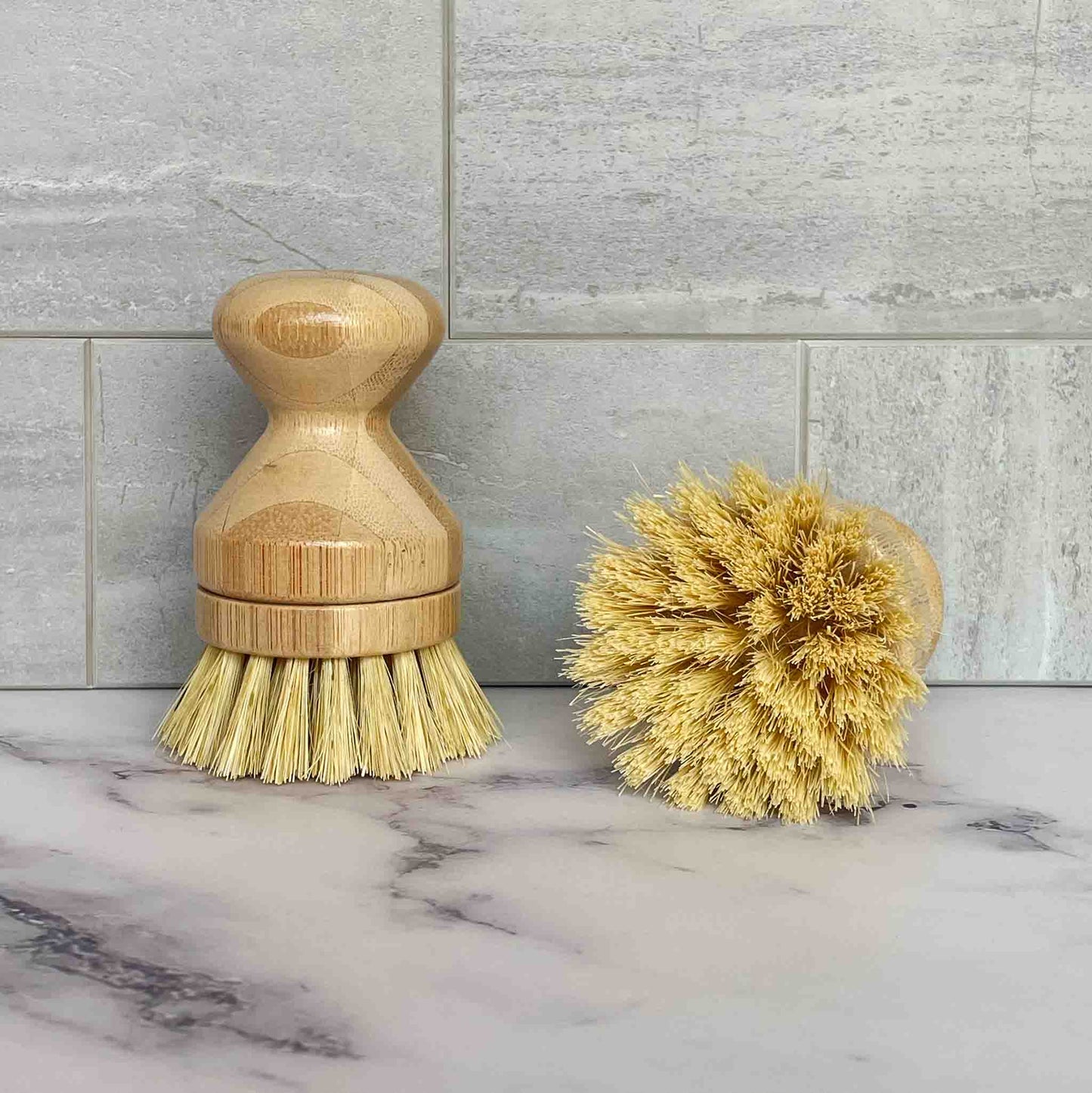 Two bamboo dish brushes with sisal bristles on a marble surface with tiled wall background
