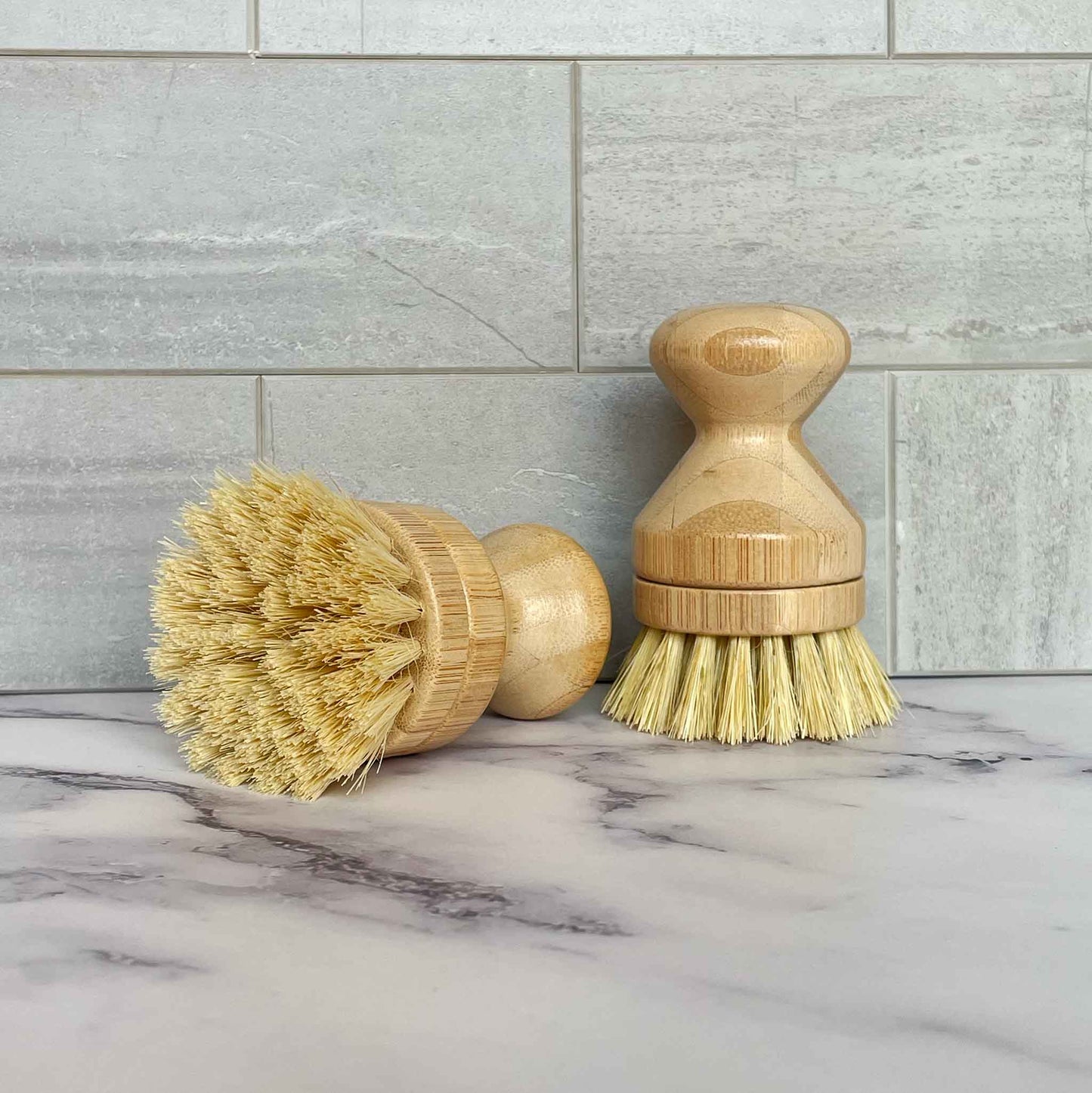 Two bamboo dish brushes with sisal bristles on a marble surface with a tiled wall background.