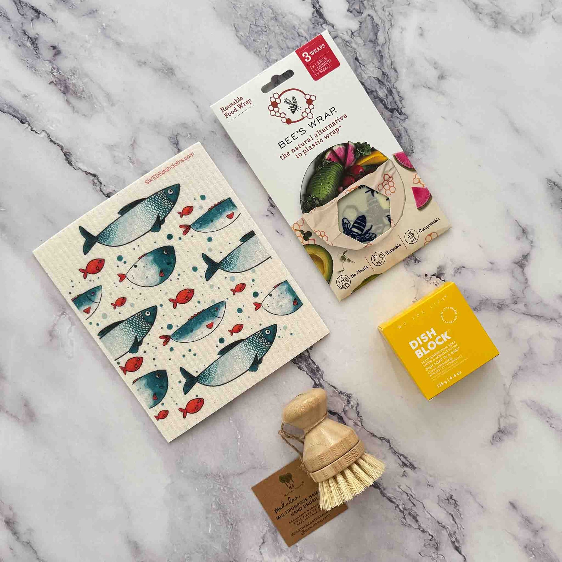 Set of eco-friendly cleaning products including a scrubber, dish block, Swedish dishcloth and Beeswax Food Wrap on a marble surface.