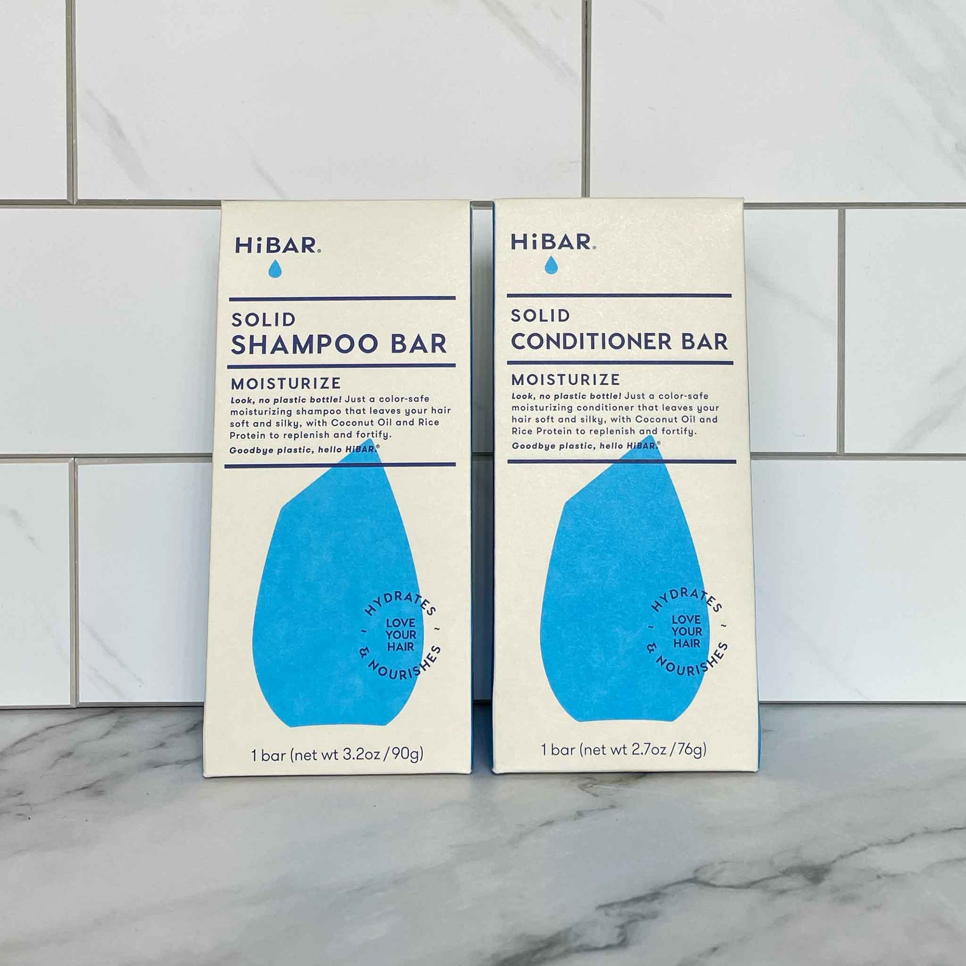 Two HiBAR Moisturize solid shampoo and conditioner bar packages on a tiled bathroom wall.