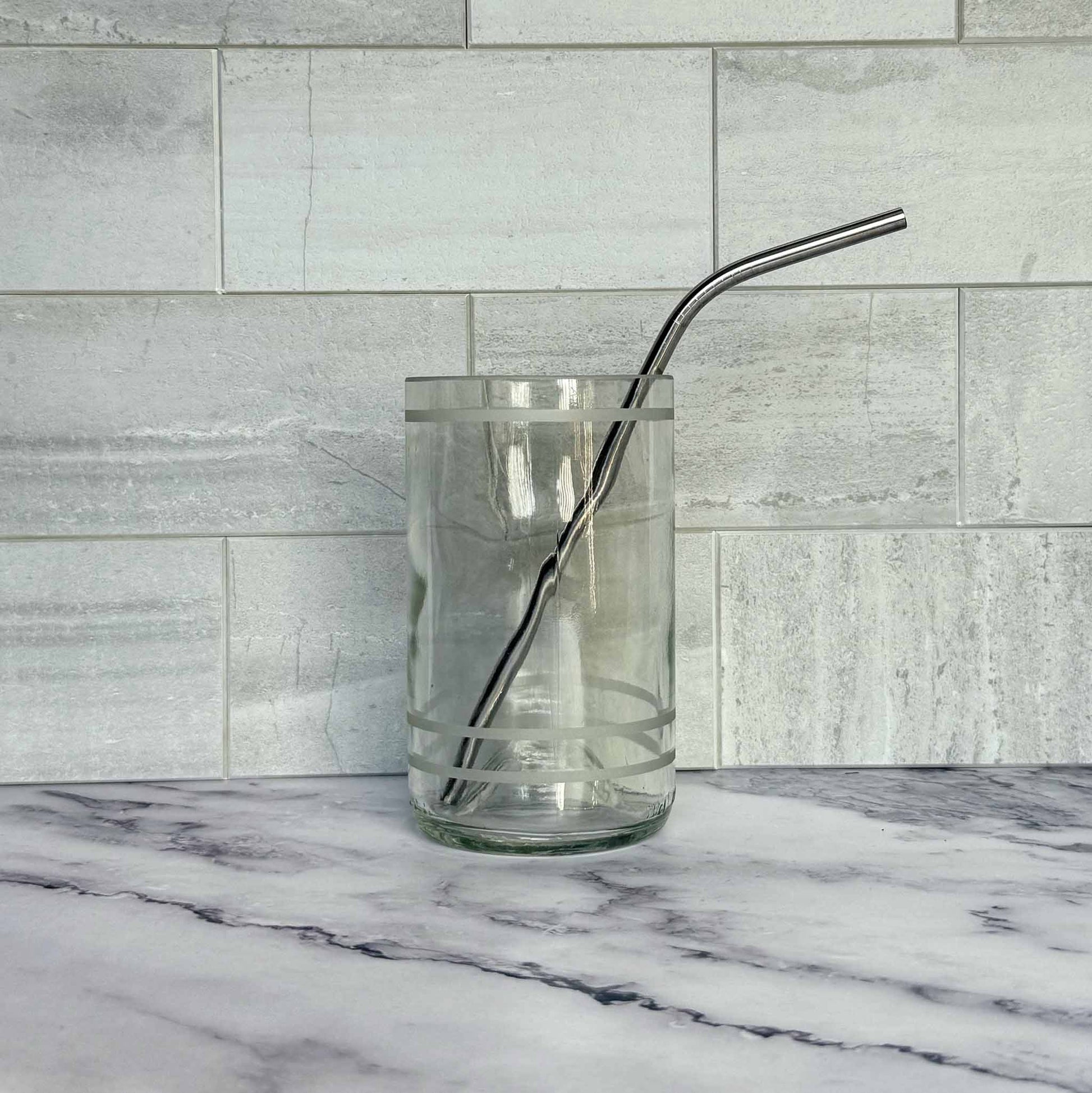 Clear glass with a stainless steel bent straw on a marble surface with a tiled wall background