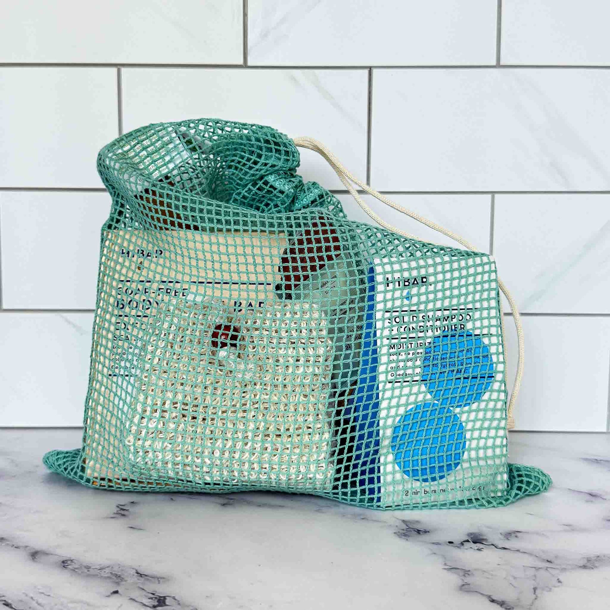 Green mesh bag with our travel kit eco-friendly products on a marble surface and tiled wall background