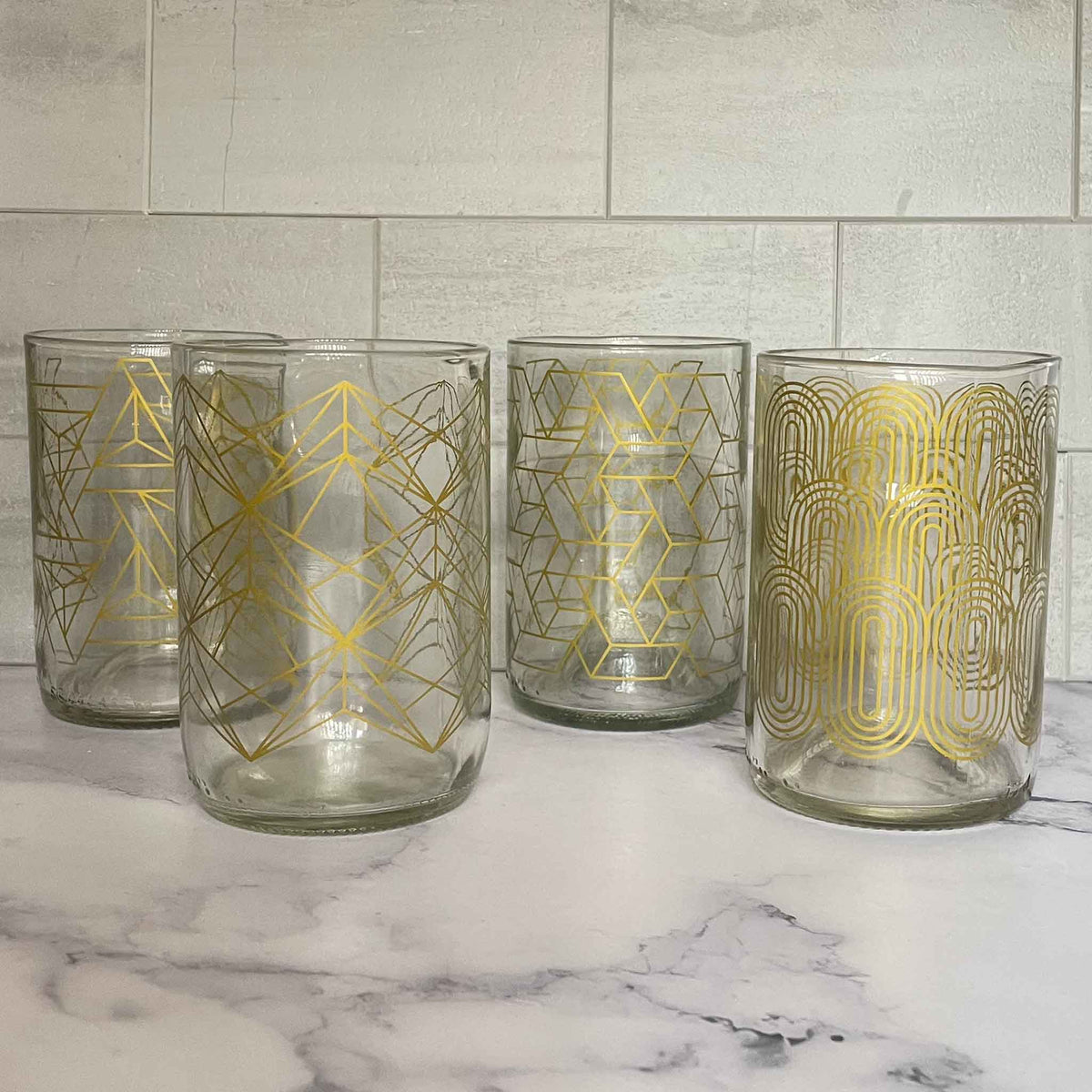 Four clear glass tumblers with gold geometric and Art Deco style patterns, arranged on a white marble surface against a light gray tiled wall.
