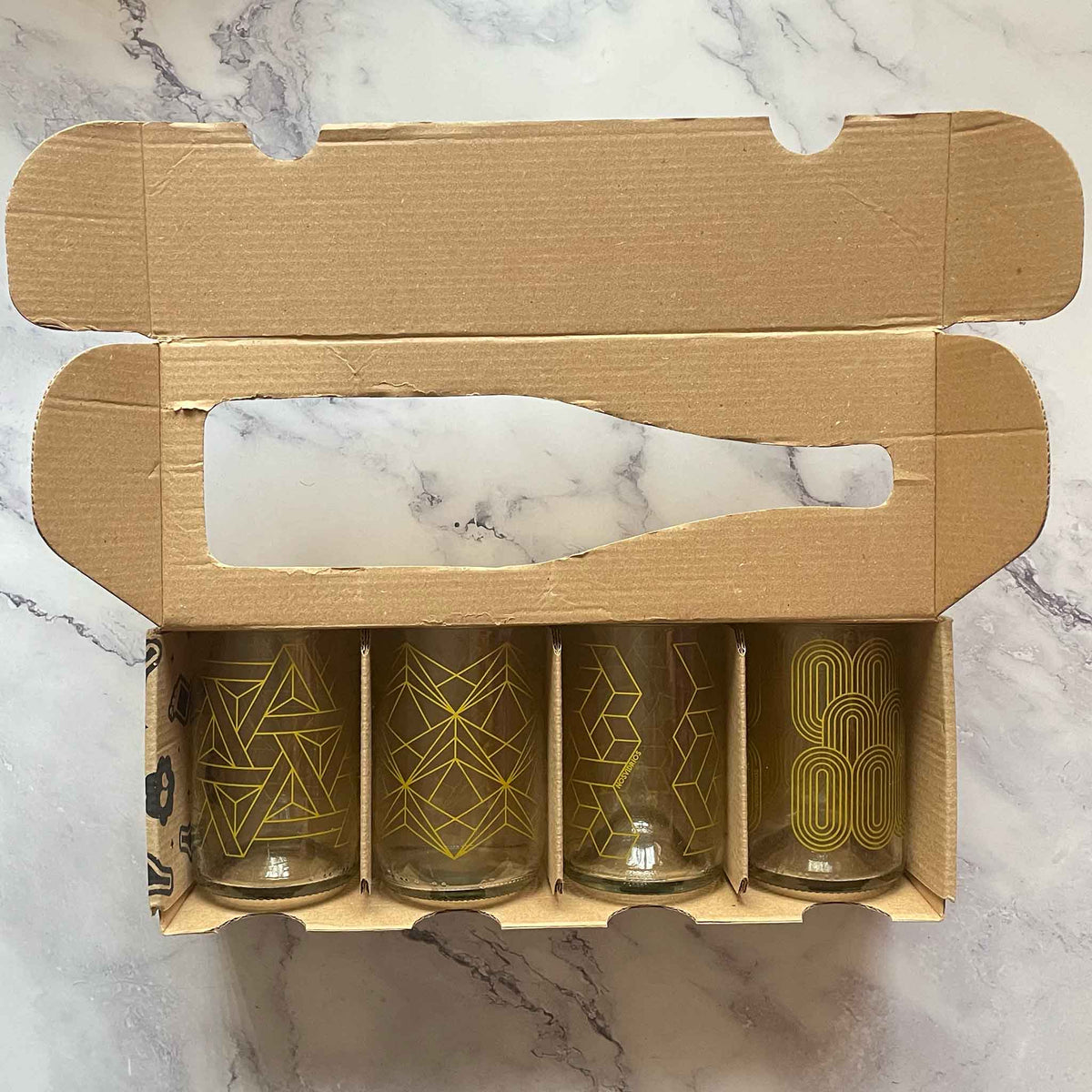 A set of four clear glasses with gold geometric line patterns, presented in an open brown cardboard box on a white marble surface.