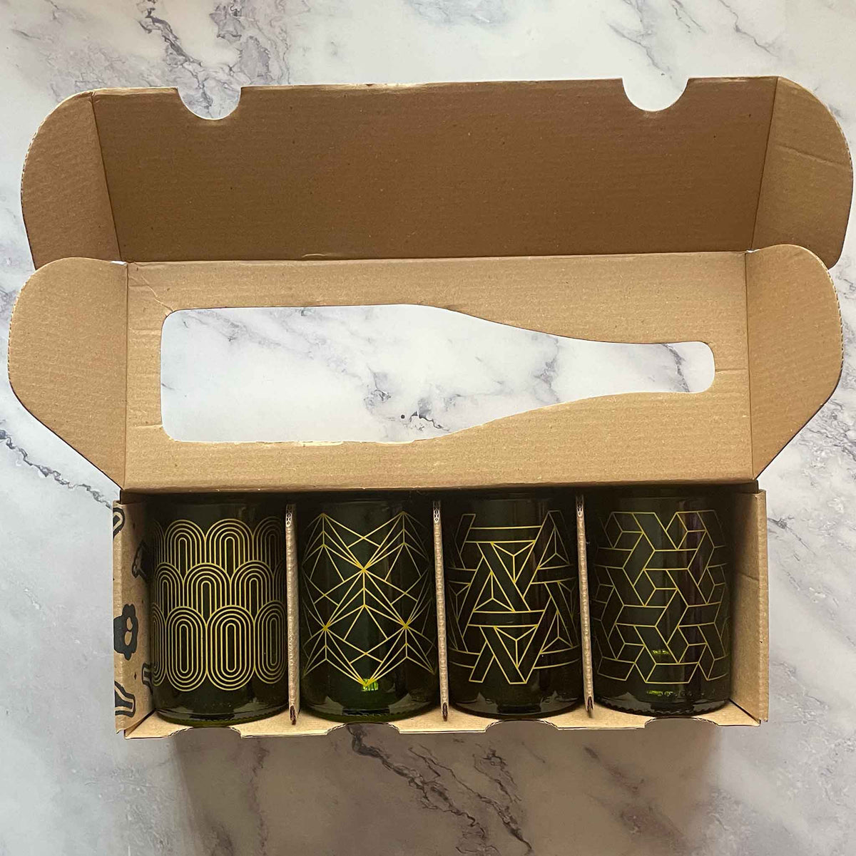 A set of four dark green glasses with gold geometric patterns, presented inside an open brown cardboard gift box on a white marble surface.
