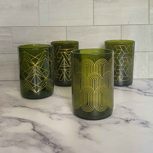 Four green glasses with gold geometric patterns arranged on a white marble surface against a light gray tiled backsplash.